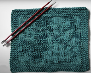 A teal knitted dishcloth with a basketweave pattern and two red knitting needles resting on it.