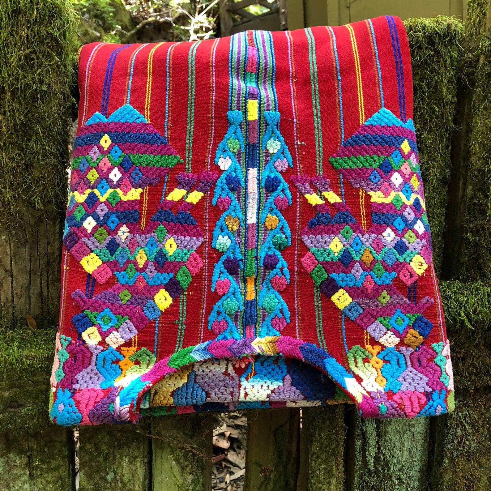 A vibrant woven textile with intricate geometric patterns in various colours the main being red, displayed against a mossy wooden background.